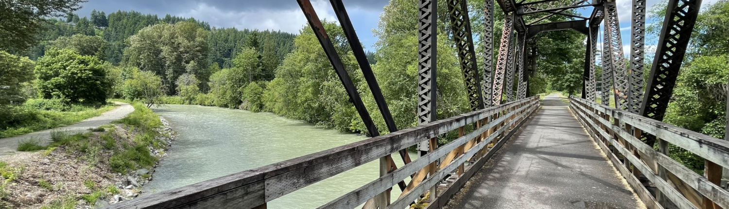 A metal frame bridge that was once a railroad line but is now a paved trail crosses the green water of the Carbon River.