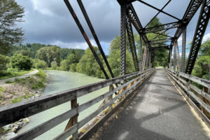 A metal frame bridge that was once a railroad line but is now a paved trail crosses the green water of the Carbon River.
