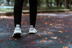 Someone in white shoes and black jogging pants stands on a paved trail covered with scattered orange leaves.